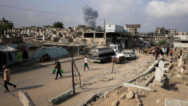 Palestinians walk past damaged graves as smoke rises following an Israeli strike, in Gaza City
