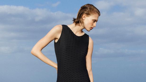 Eine Frau in einem schwarzen Kleid und Sandalen steht vor dem Meer.