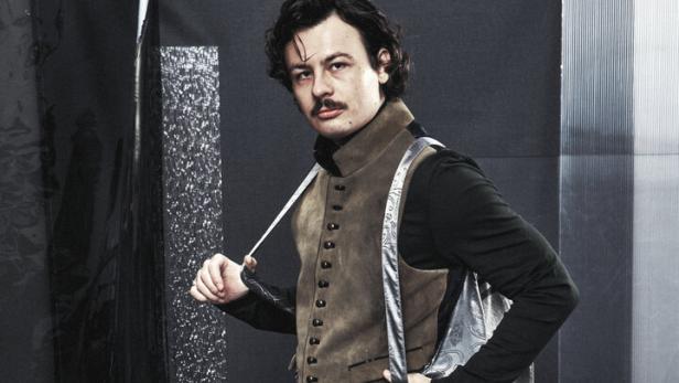 Ein Mann mit Schnurrbart trägt eine Lederhose, eine Weste und Stiefel vor einem grauen Hintergrund.