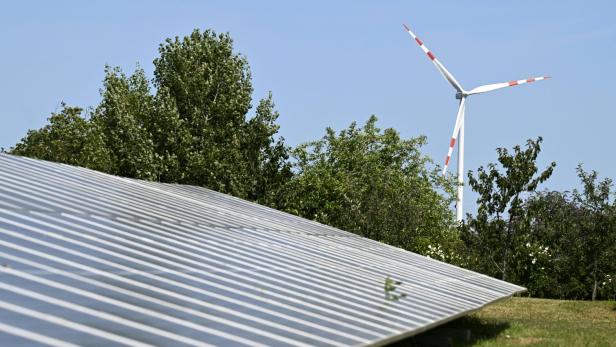 Solarmodule auf einer Wiese mit Bäumen im Hintergrund, dahinter steht ein Windrad vor blauem Himmel.