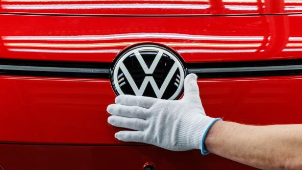 Nahaufnahme auf das VW Logo eines roten Fahrzeugs, daneben eine behandschuhte Hand