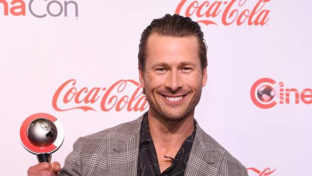 Schauspieler Glen Powell lächelt in die Kamera. Er hält einen Preis in der Hand.