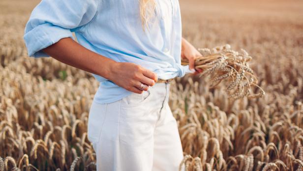 Junge Frau, die in einem Sommerfeld geht und Weizen pflückt, trägt ein Leinenhemd und Jeanshosen. Natürliche, stilvolle Kleidung.