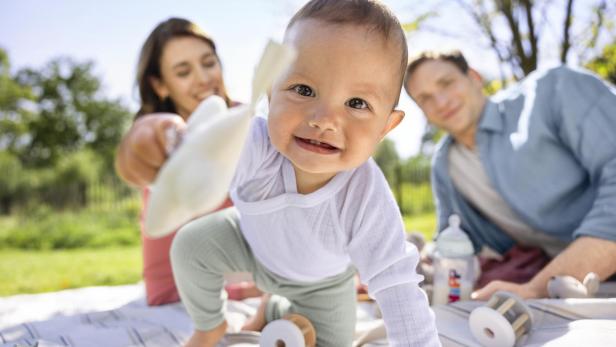 Eine Familie sitzt in einem Park auf einer Decke. Das Baby spielt mit der Kamera. 