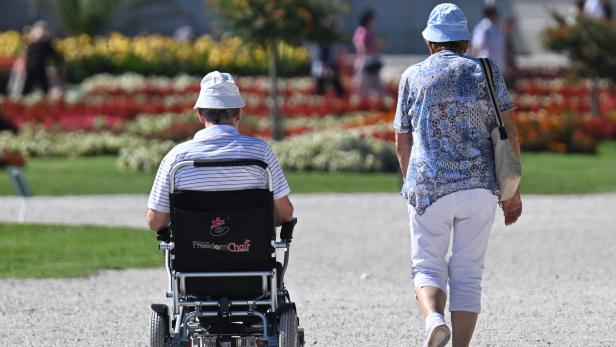 Zwei Personen, eine im Rollstuhl, bei sommerlichem Wetter auf dem Weg durch einen Park