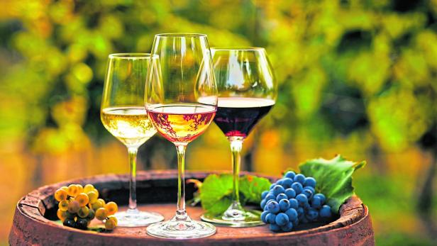 Drei Gläser Weiß-, Rosé- und Rotwein stehen auf einem alten Holzfass mit Weintrauben.