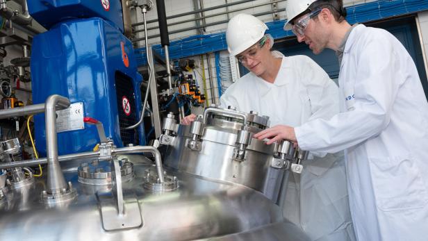 Zwei Personen in Laborkitteln und Schutzhelmen arbeiten an einem großen, silbernen Tank.