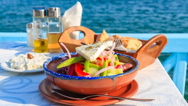 Ein griechischer Salat und Tsatsiki stehen auf einem Tisch einer Taverne in Griechenland mit Blick auf das Meer.