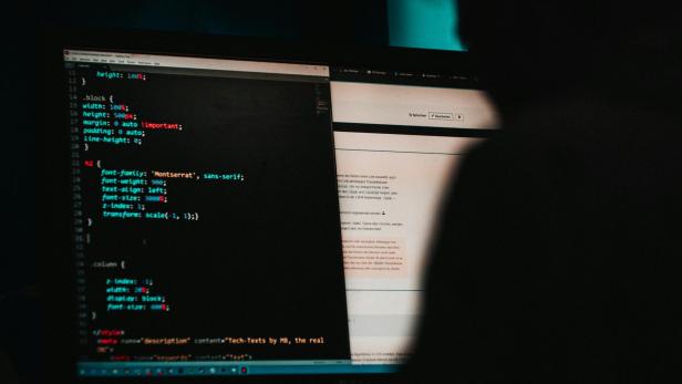 Auf einem dunklen Bild sieht man Bildschirme mit Code, davor sitzt eine dunkel gekleidete Person 