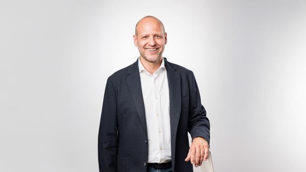 Ein Mann steht mit Sakko und weißem Hemd vor einem grauen Hintergrund und schaut in die Kamera.