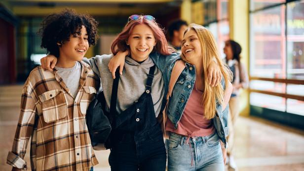 Happy high school friends walking embraced through a hallway. Drei Jugendliche schlendern Arm in Arm durch einen Schulgang