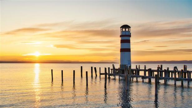 Ein rot-weißer Leuchtturm am Ufer des Neusiedlersees im Sonnenuntergang.