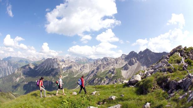 Drei Wanderer gehen auf einem grasbewachsenen Bergrücken mit Blick auf eine Bergkette.