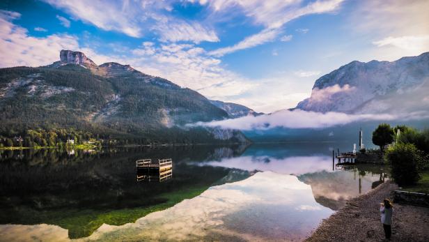 Breathtaking morning view of a woman standing on the shore of Altausseer See (Lake Aussee), Styria, Austria, drinking coffee and looking at the lake Breathtaking morning view of a woman standing on the shore of Altausseer See (Lake Aussee), Styria, Austria, drinking coffee and looking at the lake