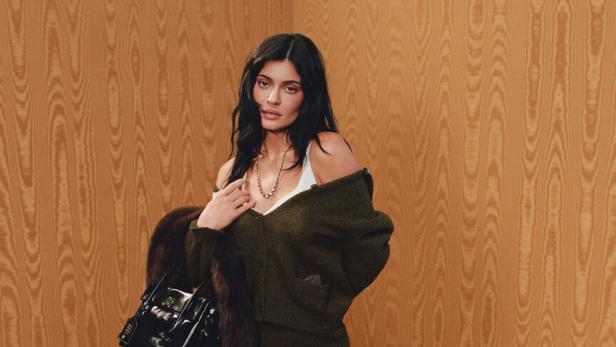Kylie Jenner posiert für Miu Miu in einem grünen Off-Shoulder-Cardigan und einem passenden Rock dazu. Außerdem trägt sie Kniestrümpfe und schwarze Schuhe zum Schnürren. Ihre Accessoires sind eine goldene Kette, eine kleine Ledertasche und ein brauner Pelzmantel.