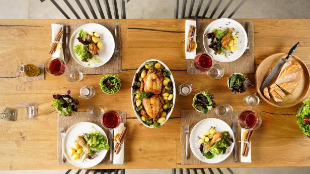 Ein üppig gedeckter Tisch: Auf vier Tellern liegen Hühnerkeulen und Gemüse. In der Mitte steht eine Schüssel mit zwei Brathühnern und Kartoffeln. Dazu viel Wein.