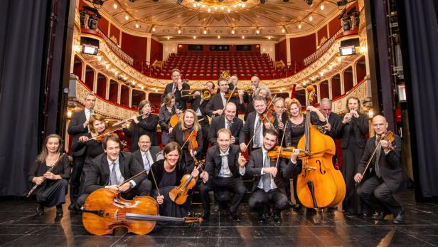 Musikerinnen und Musiker in schwarzer Kleidung stehen auf der Bühne Baden, im Hintergrund die hell erleuchtete Zuschauerränge.