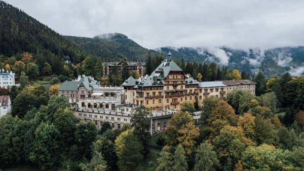 Das große Südbahnhotel mit seinen verschiedenen Trakten und großem Balkon. Es thront auf einem bewaldeten Hang. Im Hintergrund erheben sich Berge, Nebel lichtet sich