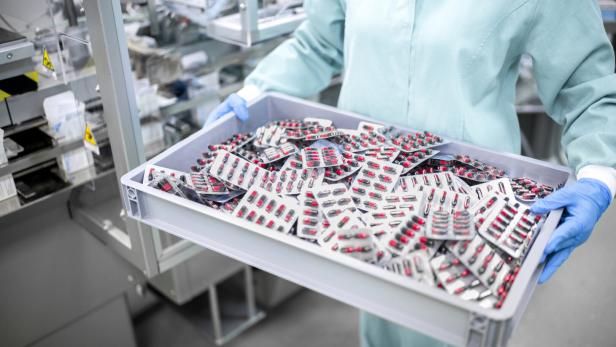 A worker in protective gear carefully carries a container filled with blister-packaged capsules Frau mit Handschuhen trägt Tablett mit Tabletten-Packungen