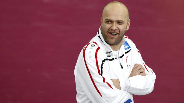 Ein Mann in einer weißen Trainingsjacke lächelt vor einem roten Hintergrund.