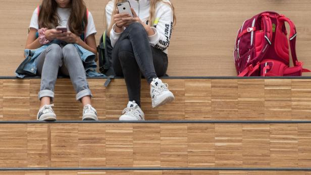 Zwei Mädchen sitzen auf einer Treppe und schauen auf ihre Smartphones. Ein Rucksack steht daneben.