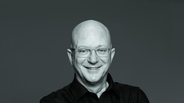 Niki Glattauer Ein älterer Mann mit Glatze, Brille und schwarzem Hemd lächelt mit verschränkten Armen vor neutralem Hintergrund.