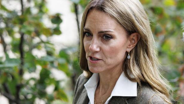 Prinzessin Kate Prinzessin Kate steht draußen vor grünem Laub. Sie trägt ein Sakko mit weißem Hemd und Ohrringe.