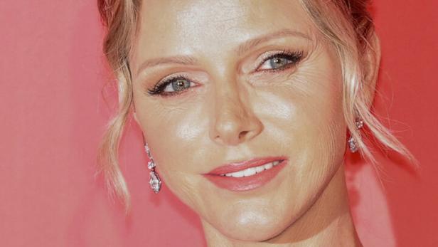 Fürstin Charlène von Monaco trägt ein blaues Kleid und eine funkelnde Halskette vor einem roten Hintergrund.