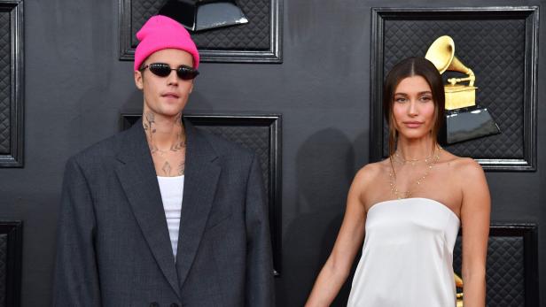 Justin und Hailey Bieber halten Händchen vor einer dunklen Wand mit goldenen Akzenten.
