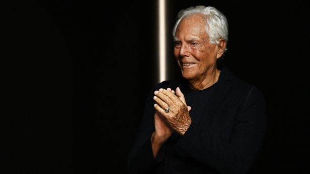 Giorgio Armani Giorgio Armani lächelt und klatscht vor dunklem Hintergrund Beifall.