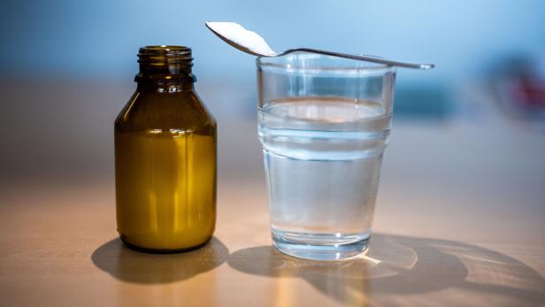 Braune Medikamentenflasche neben einem Wasserglas mit Löffel voller weißem Pulver.
