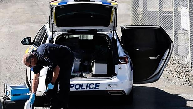 Ein Polizist mit Handschuhen untersucht Gegenstände hinter einem geöffneten Polizeiauto auf einem Schotterweg.