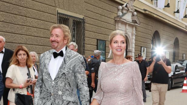 Thomas Gottschalk und Karina Mroß Thomas Gottschalk im gemusterten Sakko und Karina Mroß im Paillettenkleid gehen händchenhaltend an einem Gebäude entlang.