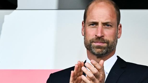Prinz William klatscht in einem dunklen Anzug vor einem hellen Hintergrund.