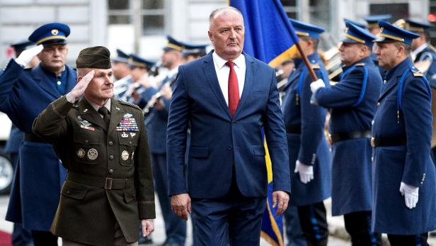Ein Mann im Anzug und ein Soldat salutieren vor einer Militärkapelle mit Flagge.