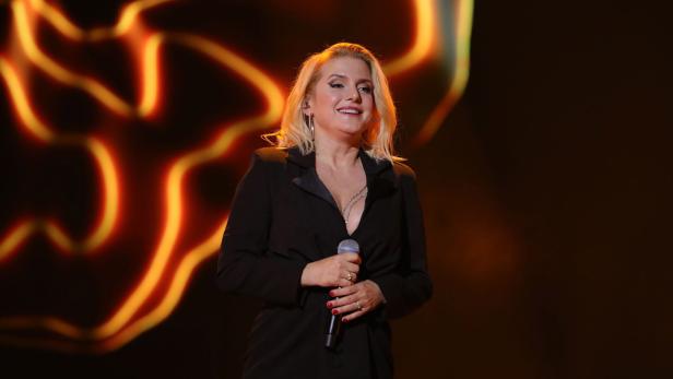 26th Annual Jose Carreras Gala Jeanette Biedermann im schwarzen Blazer hält ein Mikrofon vor einem orangefarbenen Hintergrund.