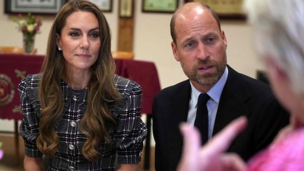 Kate und William sitzen an einem Tisch und hören einer Person aufmerksam zu.