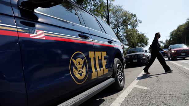 Ein dunkelblaues Fahrzeug mit ICE-Logo steht am Straßenrand, während eine Person mit Maske die Straße überquert.