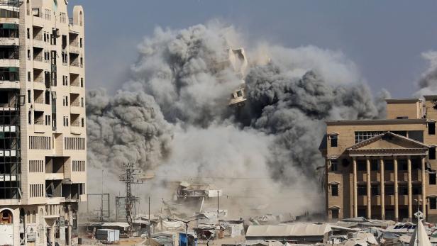 Dichte Rauchwolken steigen zwischen beschädigten Gebäuden und Zelten auf, während im Hintergrund eine Explosion stattfindet.