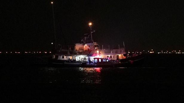 Ein brennendes Schiff liegt nachts auf dem Wasser, umgeben von Dunkelheit und fernen Lichtern am Horizont.