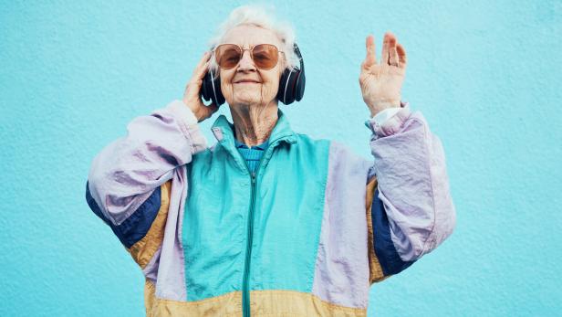 Happy, elderly and woman with headphone music for wellness, fun and retirement leisure. Urban, retro and funky senior person listening to feel good streaming on headphones at blue wall. Eine ältere Frau mit Sonnenbrille und Kopfhörern tanzt vor einem hellblauen Hintergrund.