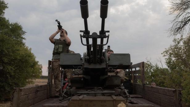Zwei Soldaten stehen auf einem Militärfahrzeug mit einer großen Flugabwehrkanone, einer blickt durch ein Fernglas in den Himmel.