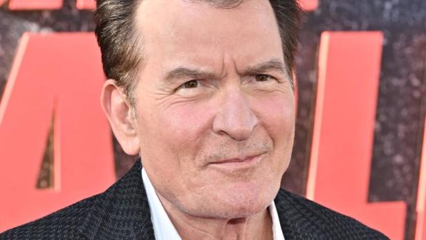 Charlie Sheen Charlie Sheen lächelt in einem weißen Hemd und einem dunklen Sakko vor einem roten Hintergrund.