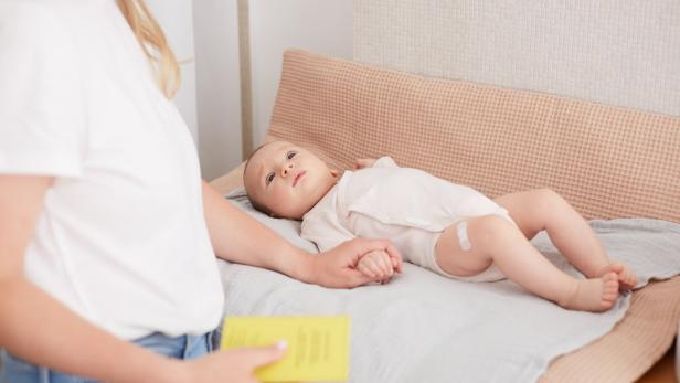 Welt-Sepsis-Tag 2025: Babys, Kleinkinder und Jugendliche bestmöglich vor Meningokokken-Erkrankungen schützen Ein Baby liegt auf einer weichen Unterlage, hält die Hand einer erwachsenen Person und hat ein Pflaster am Oberschenkel.