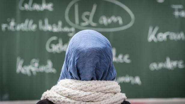 Eine Person mit blauem Kopftuch und weißem Schal steht vor einer Tafel mit islambezogenen Begriffen.