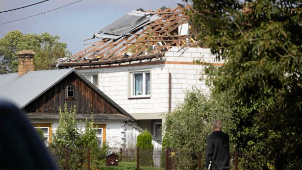 Ein Haus mit stark beschädigtem Dach, bei dem große Teile der Dacheindeckung fehlen, steht inmitten von Bäumen und Sträuchern.