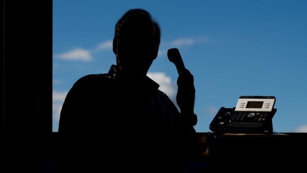 Betrüger prellen Senioren Eine Silhouette einer Person telefoniert mit einem Tischtelefon vor einem Fenster mit blauem Himmel.