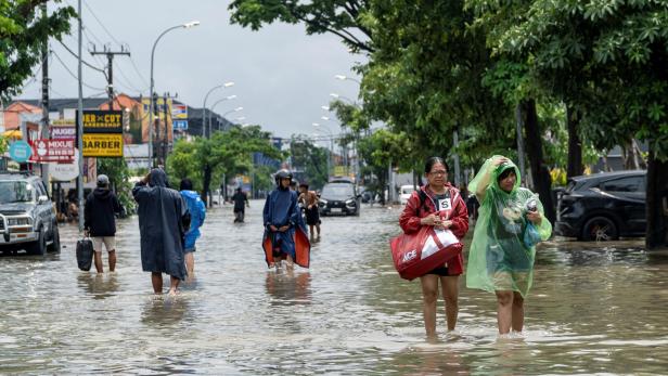 Menschen waten mit Regenmänteln und Taschen durch eine überflutete Straße, während Autos im Wasser stehen.
