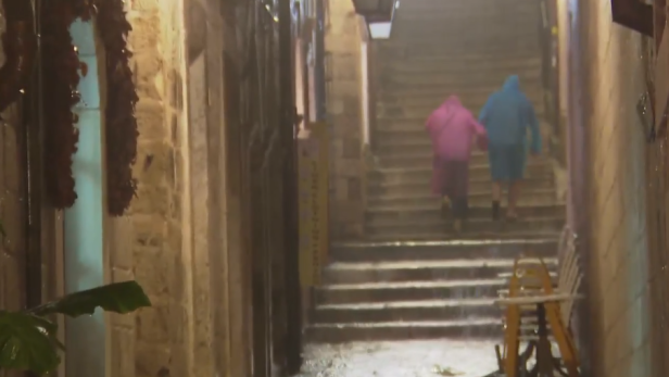 Zwei Personen in Regenmänteln steigen bei starkem Regen eine nasse Steintreppe hinauf.