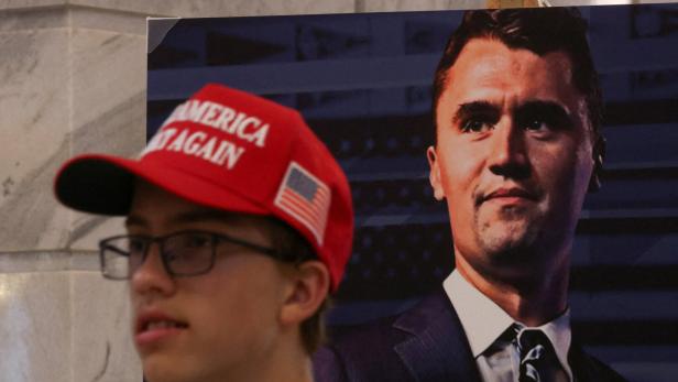 Charlie Kirk fatally shot at Utah Valley University event Ein junger Mann mit roter Kappe und Brille steht vor einem großen Porträt eines Mannes im Anzug.